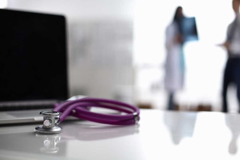  Purple stethoscope on a white desk beside a laptop, with blurred healthcare professionals in the background.