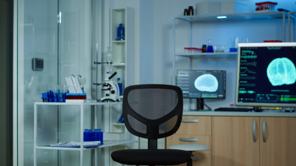  Medical imaging room with a black mesh chair, two monitors displaying brain scans, and lab shelves in the background.