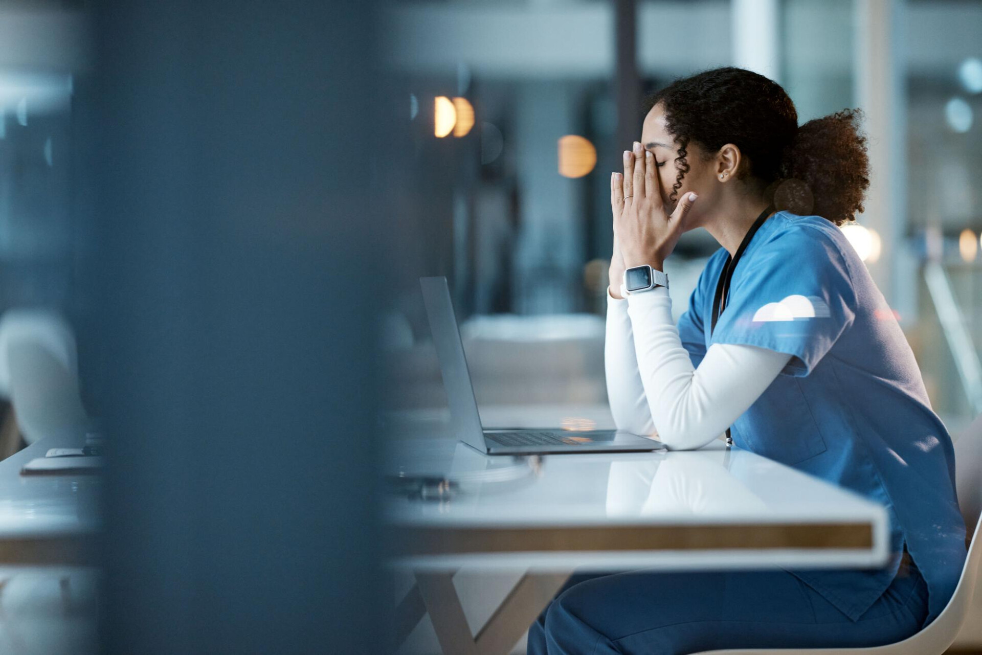  Healthcare worker in scrubs sits at a desk with a laptop, hands covering face in a stressed or exhausted pose