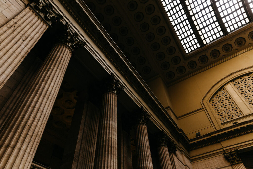 ‘Four Years for a Hearing’: Employment Tribunal Delays Exposed - Wolf Law Wolf Law 141 Interior of a grand classical hall with tall fluted columns, ornate capitals, and a coffered ceiling with a large grid skylight on the right.