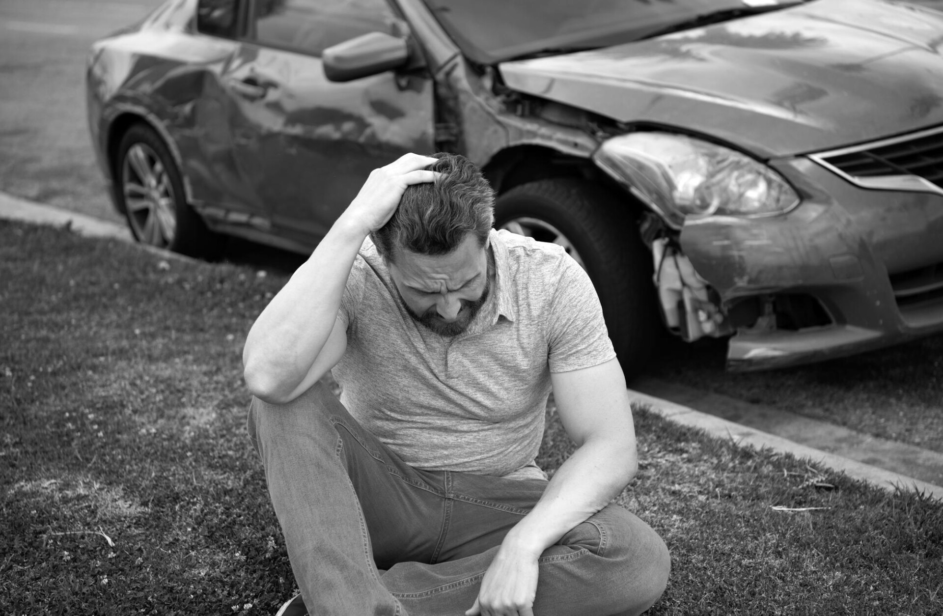 Man sitting on grass beside a crashed car, hand on head in distress after an accident.