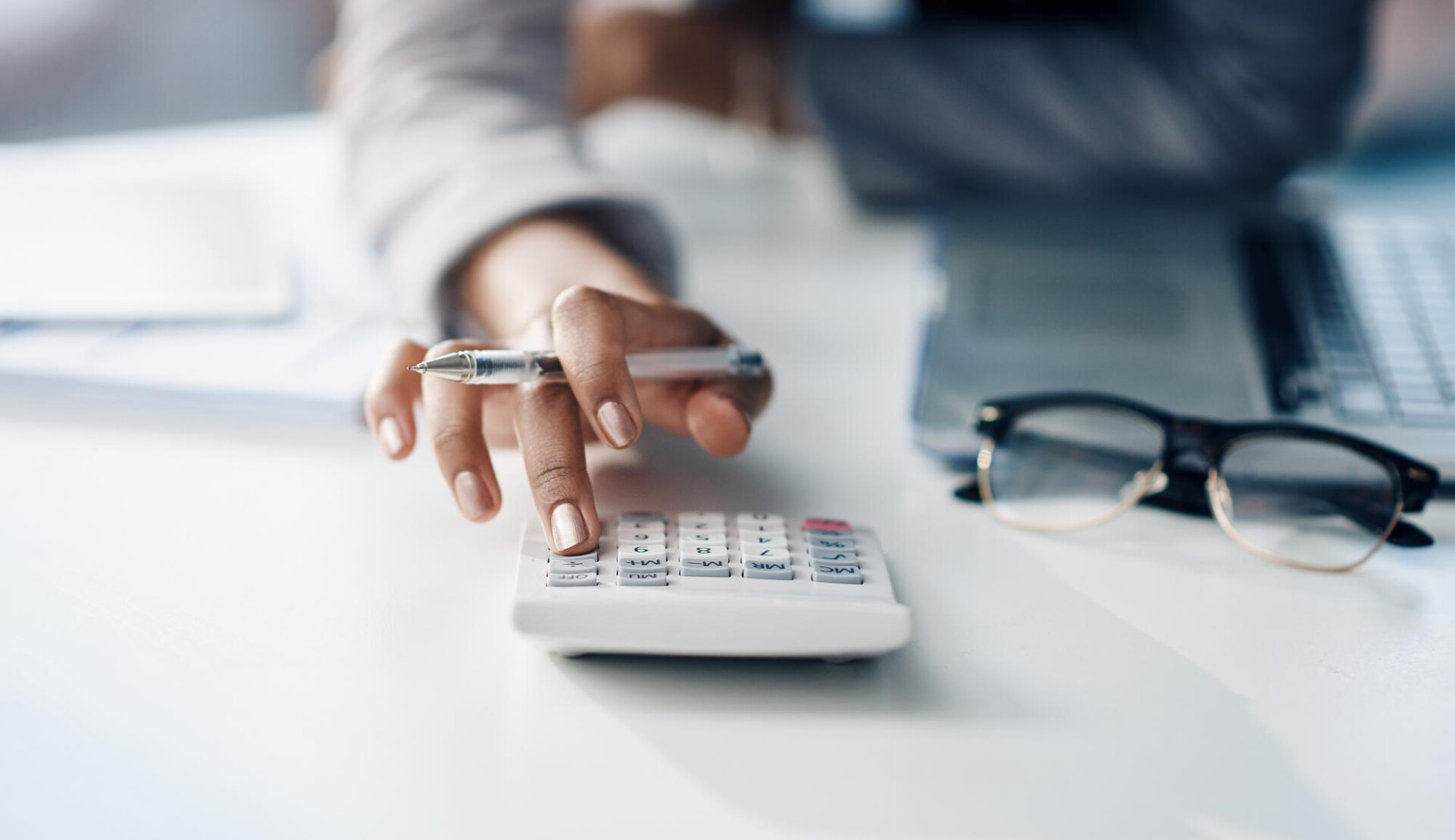 Person uses a calculator on a white desk, holding a pen and wearing rings, with a laptop and glasses in the background.