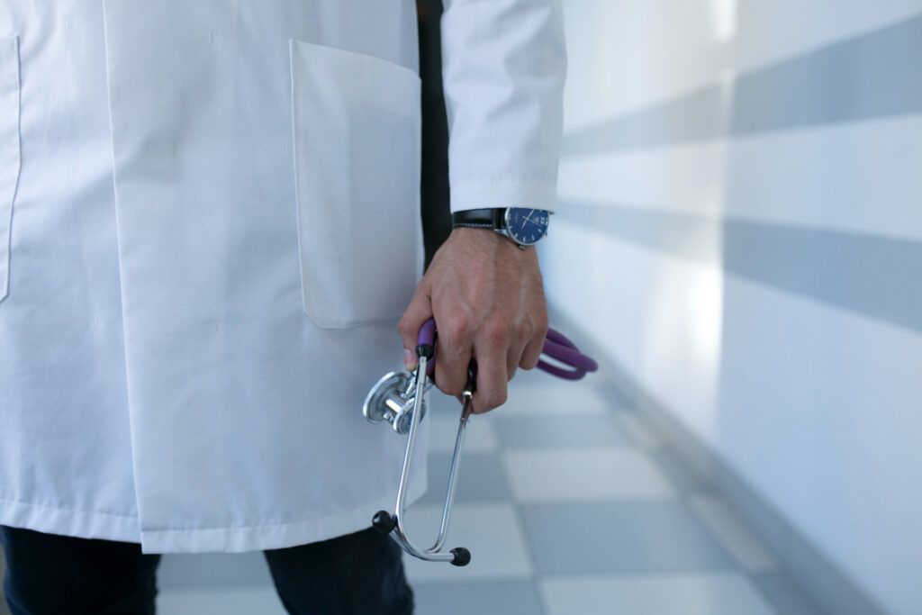 Medical professional in a white coat holding a stethoscope and medical scissors at their side, ready for examination.