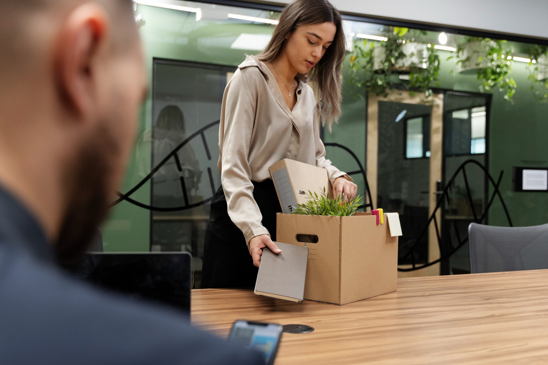 Woman packs a plant into a cardboard box at a wooden table in a modern office; a man is blurred in the foreground.