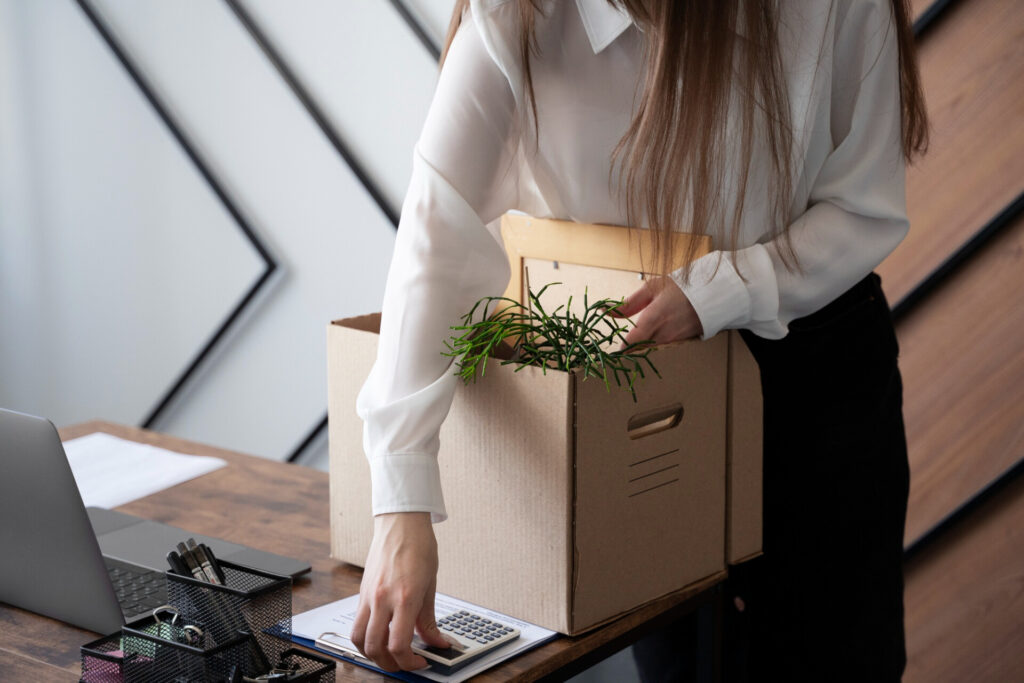 Person in a white shirt places a cardboard box with a green plant on a desk beside a laptop.