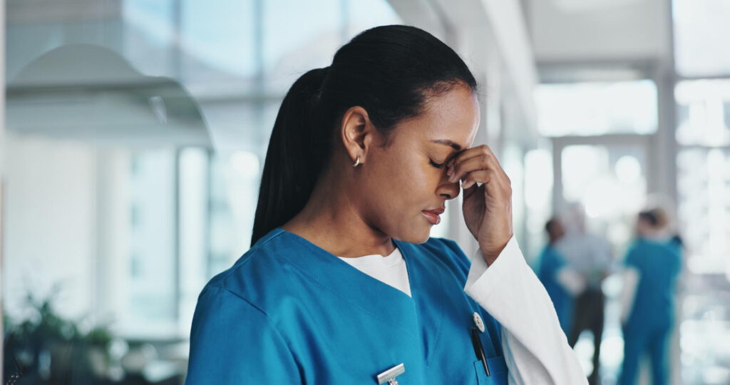 Nurse in blue scrubs in a bright hospital hallway, hand to forehead showing stress or fatigue.