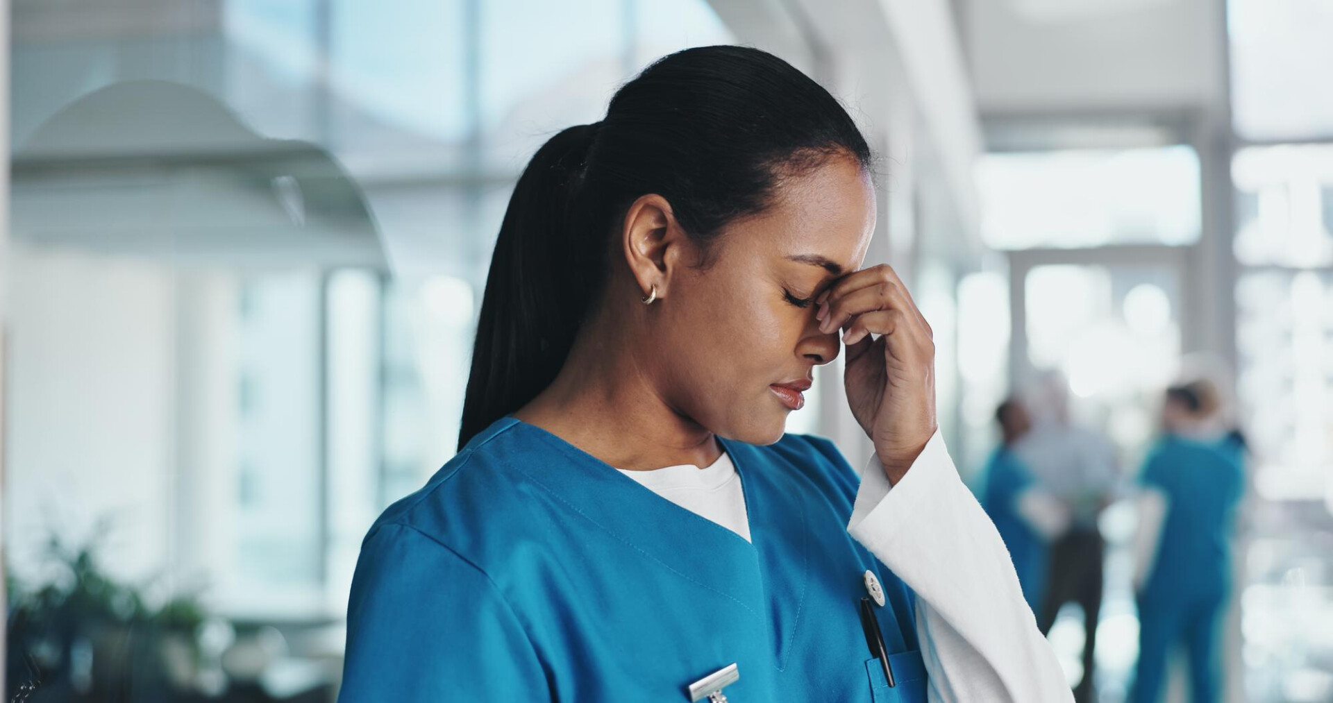 Nurse in blue scrubs in a bright hospital hallway, hand to forehead showing stress or fatigue.