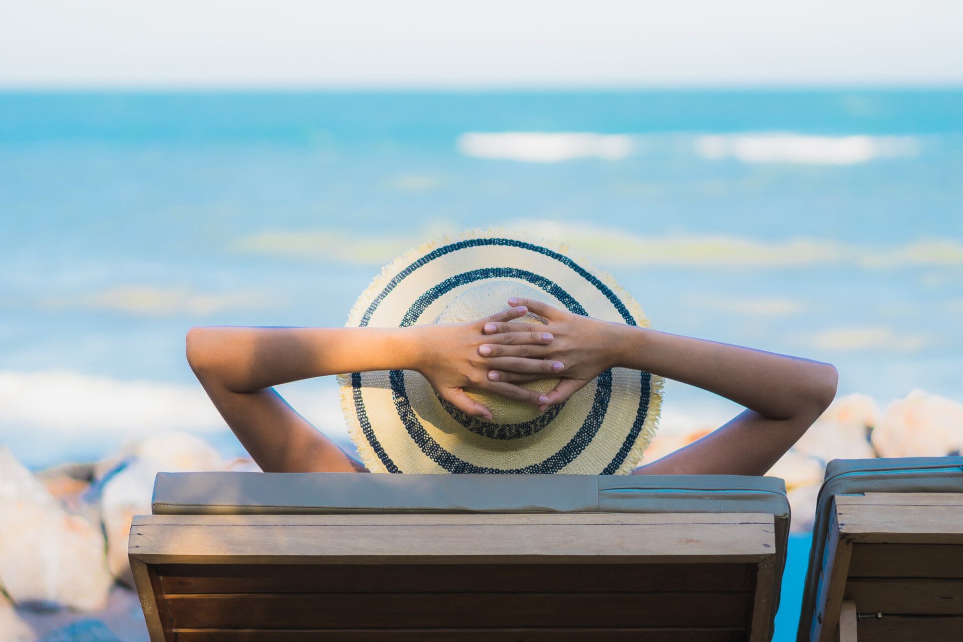 Person with a straw hat rests hands behind their head on a wooden bench at the beach, ocean visible in the background.