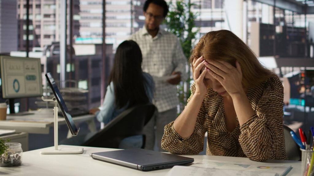 Woman sits at a desk in an office, holding her head in frustration while colleagues converse in the background.