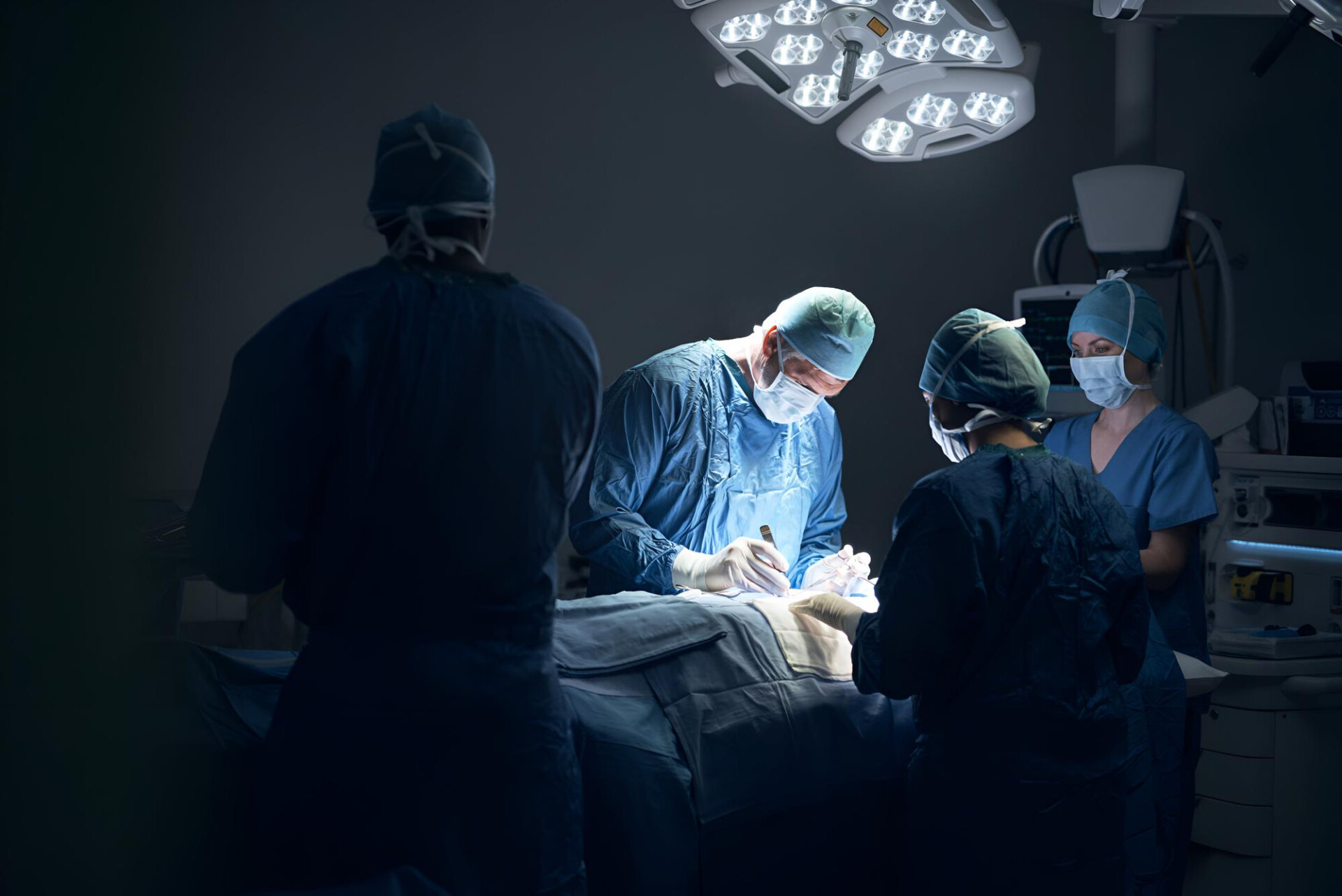Surgeons in blue scrubs and masks perform surgery under bright overhead lights around a patient on the operating table.