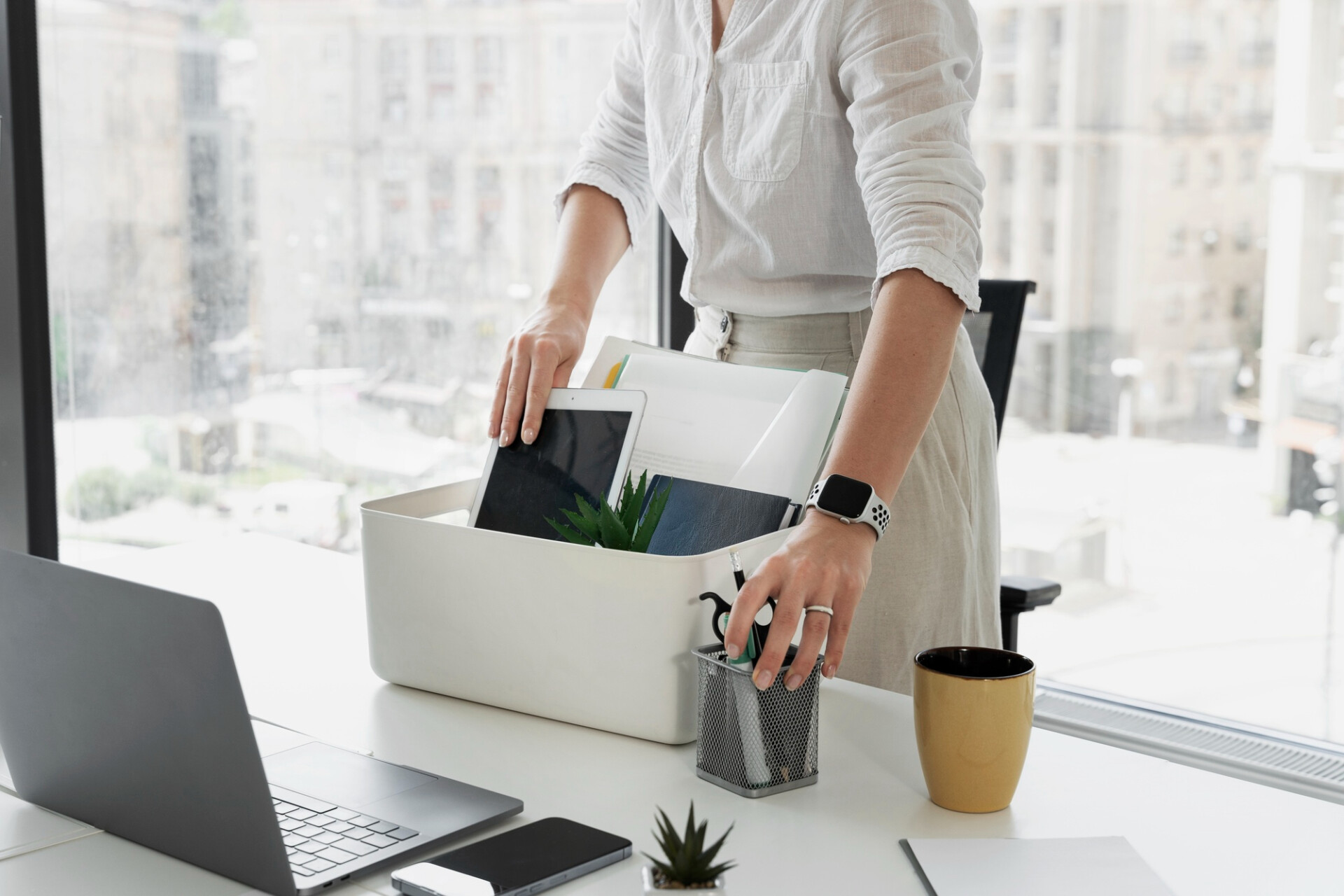 Person in a white shirt places folders and a tablet into a white desk organizer on a bright office desk.