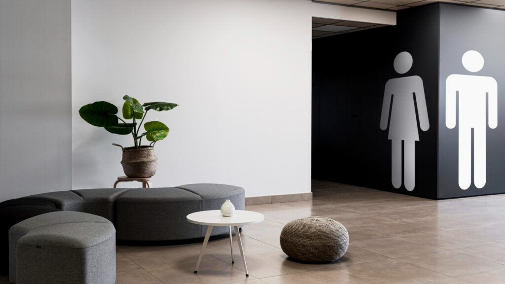 Single-Sex Facilities in the Workplace: UK Employment Law Guidance - Wolf Law Wolf Law 2149956484 Modern lounge with grey curved seating, a potted plant in a woven basket, and restroom icons on a dark wall to the right.