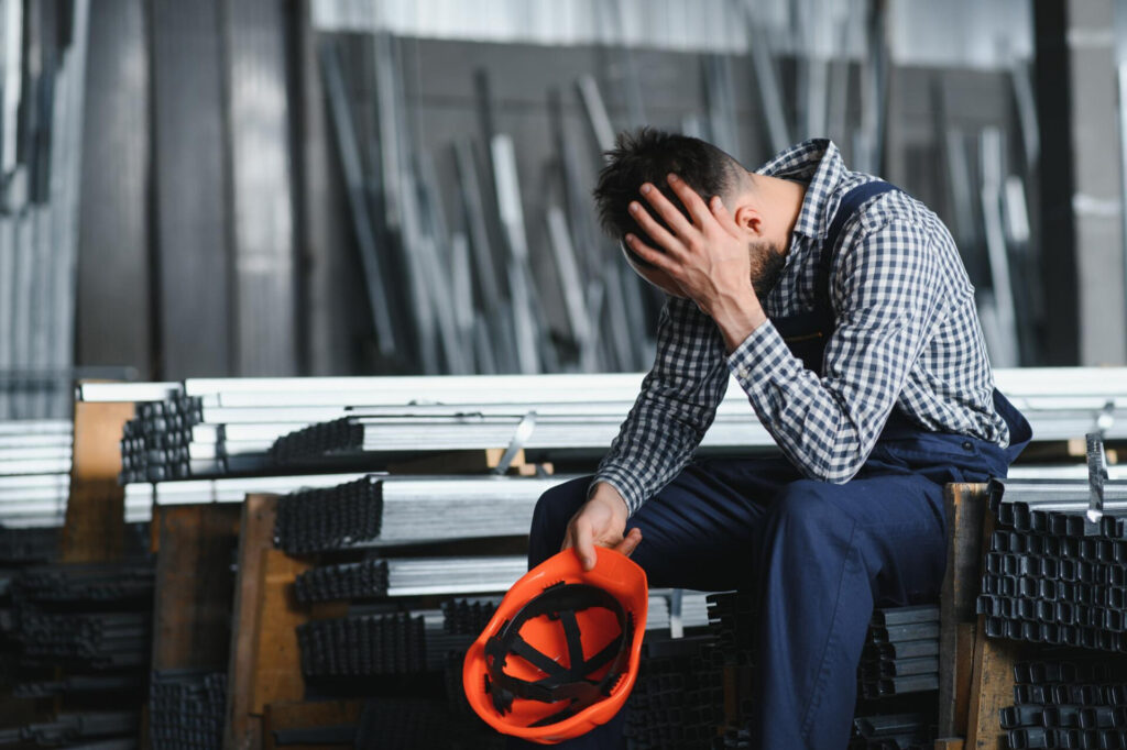 Unfair Dismissal: What You Need to Know About Your Employment Rights - Wolf Law Wolf Law 38869 Man in plaid shirt and overalls sits among stacked metal beams, head in hands, holding an orange hard hat, looking stressed in a warehouse.