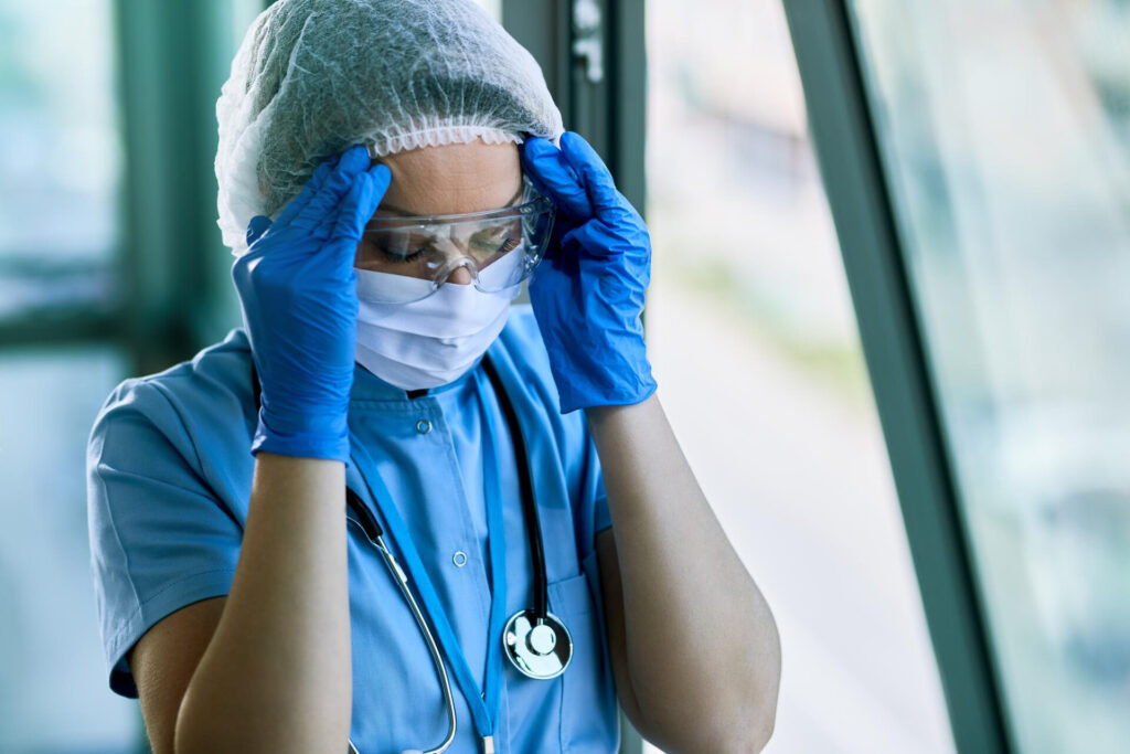 Healthcare worker in blue scrubs adjusting protective goggles while wearing a surgical cap and mask, near a windowed environment with a stethoscope around the neck