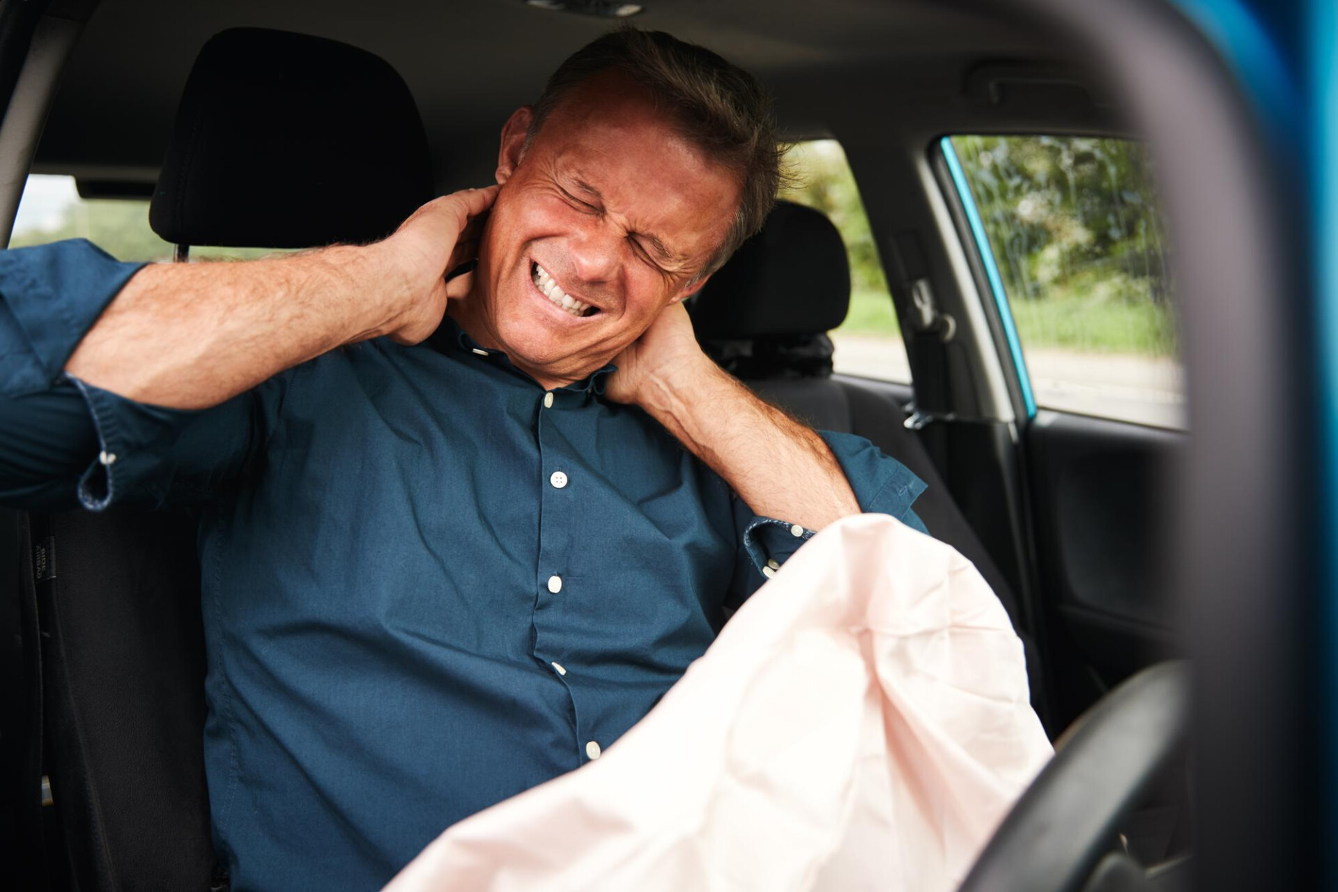 Man in a blue shirt sits in a car, gripping his neck with a pained expression as if in neck pain.