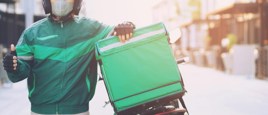 Thousands of Just Eat Couriers Launch Legal Action: What You Need to Know About Worker Rights - Wolf Law Wolf Law 6121 Delivery worker in green jacket with mask and helmet, holding a green insulated delivery bag on a scooter/bike outdoors.