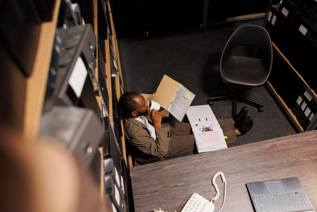Man in a suit sits on the server-room floor, drinking from a mug while reading a binder of documents amid racks and cables