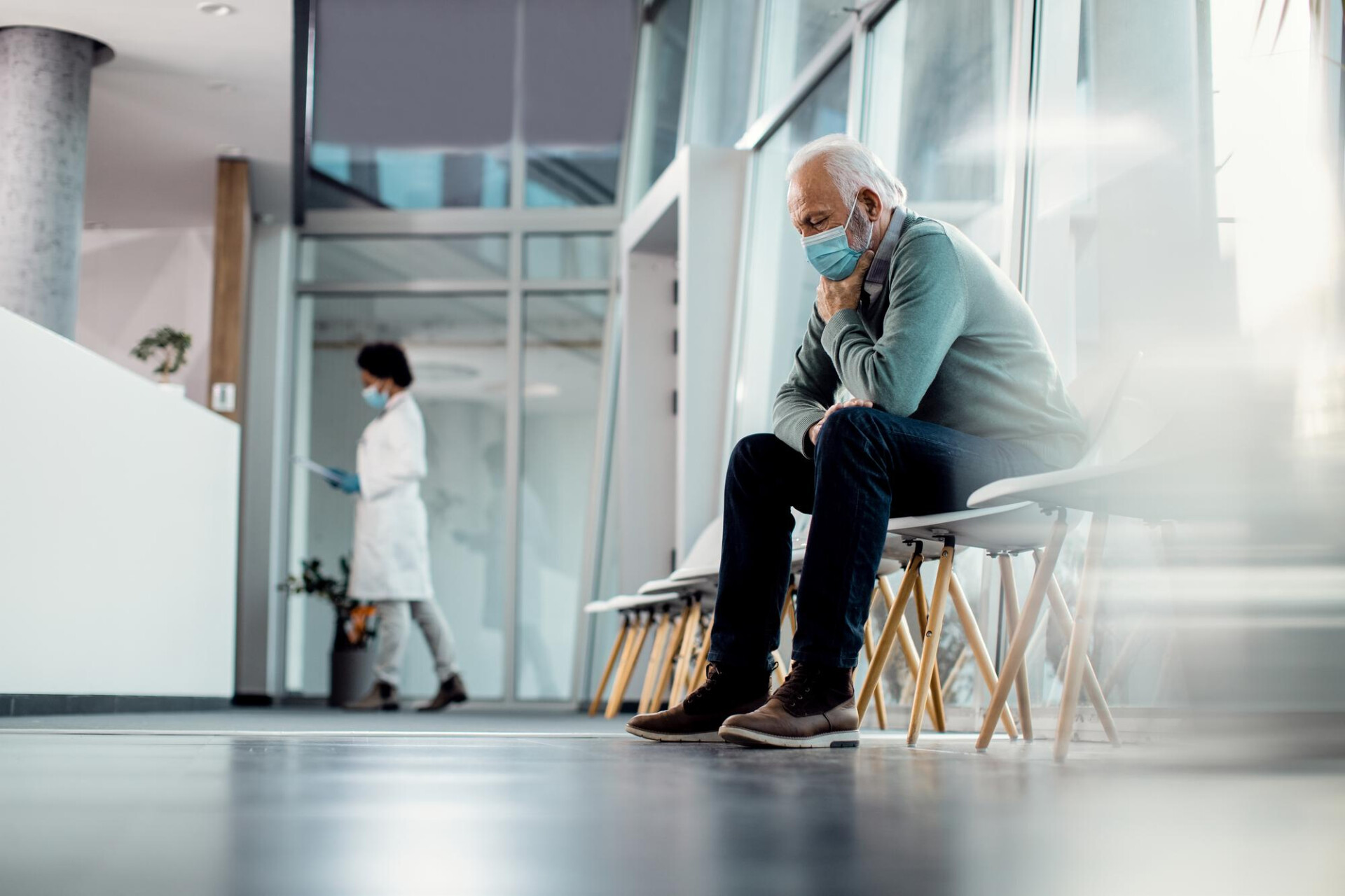 Elderly man wearing a mask sits on a row of chairs in a modern clinic lobby, nurse visible in the background.