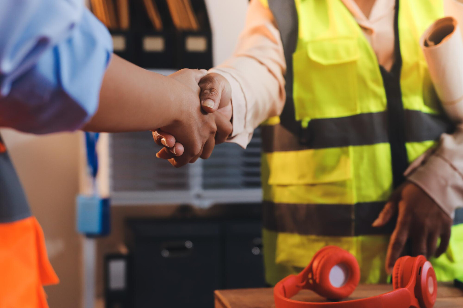 Two people shake hands in a workshop; one wears a yellow safety vest, suggesting a collaborative moment at a construction site.