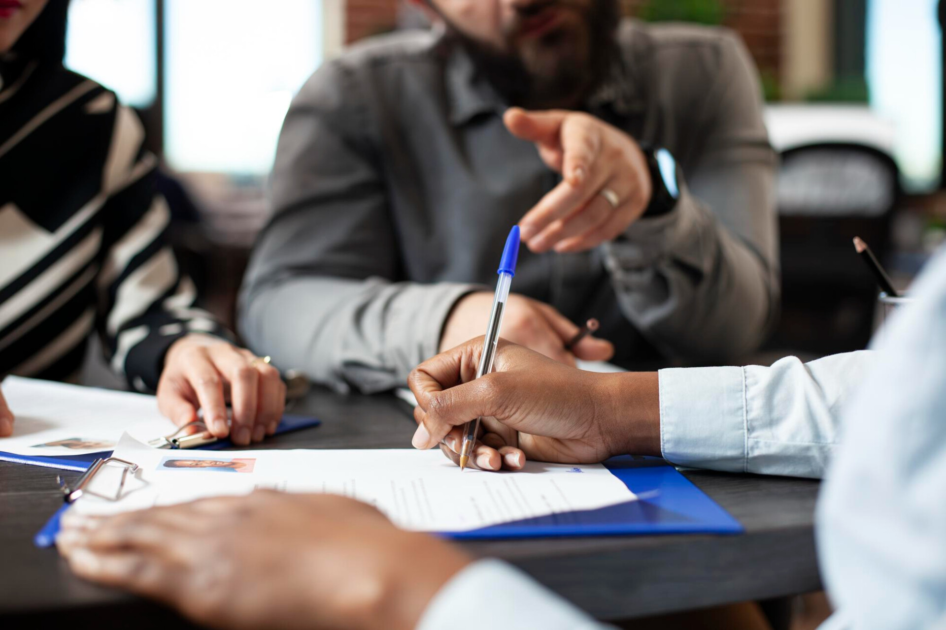 Several people sign documents at a table with pens and a binder visible, in a professional meeting setting.