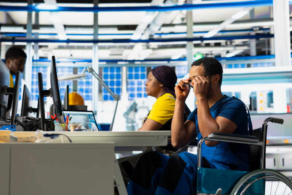 Employment Law | Expert Legal Representation | Wolf Law Wolf Law Constructive Dismissal and Disability: What Employers Need to Know - Wolf Law Wolf Law 125819 Man in a wheelchair sits at a desk with multiple computer monitors in a bright lab or office space as colleagues work nearby.