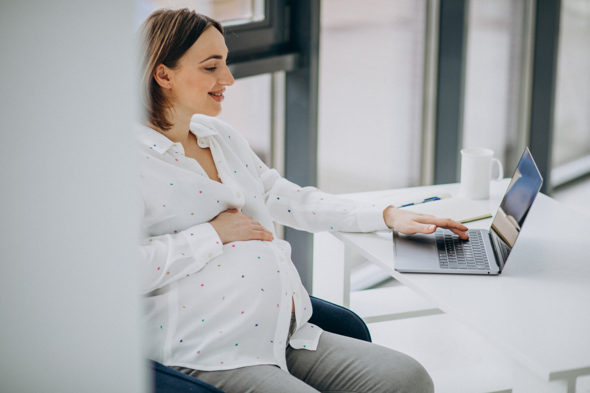 Pregnant woman in a white dotted blouse sits at a desk using a laptop by a window.