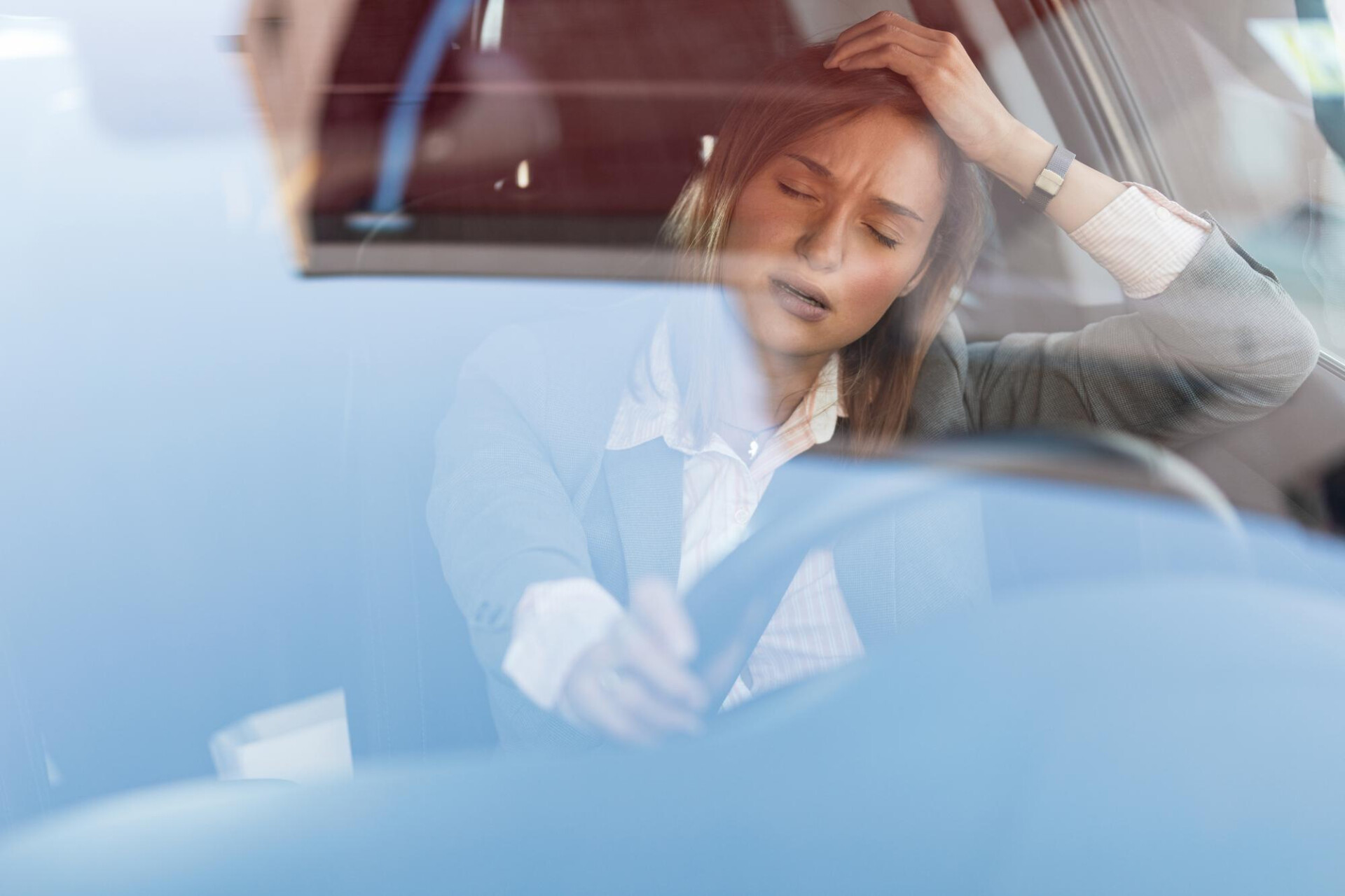 Woman in a blazer inside a car, eyes closed, hand pressed to her head in apparent stress or headache.