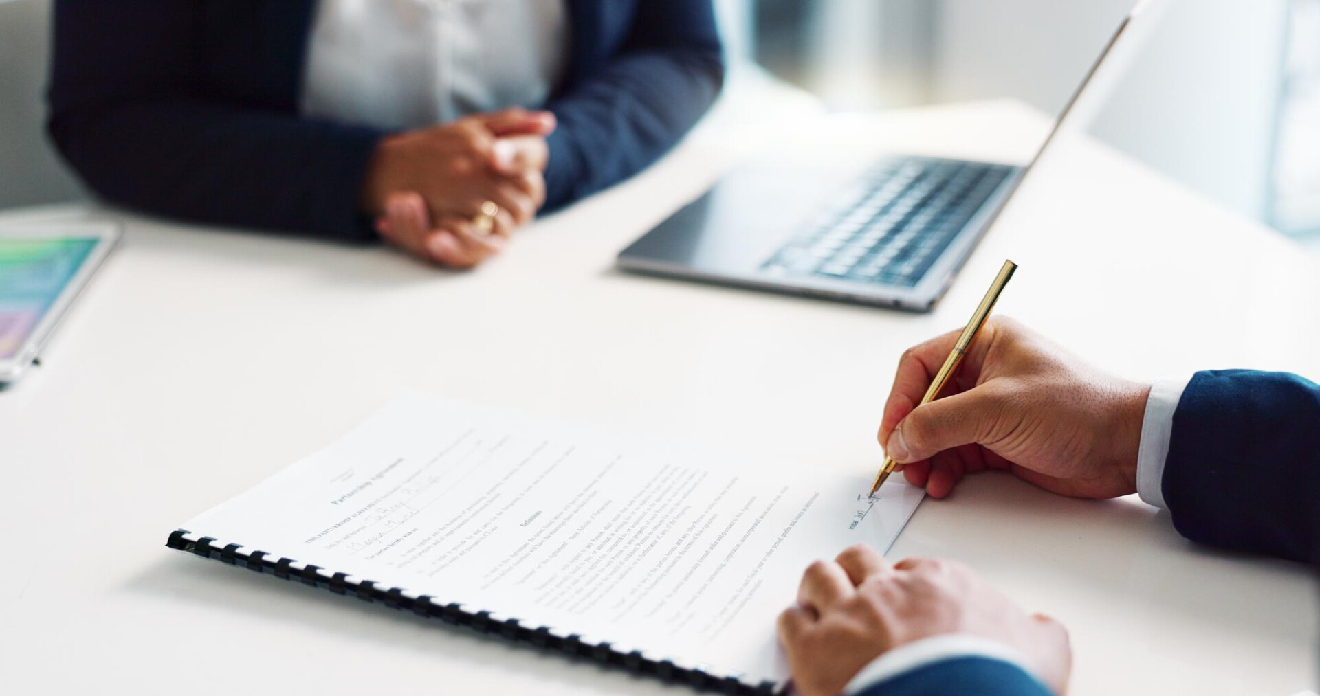 Two professionals sign a document on a white table, with a laptop nearby in a bright office.
