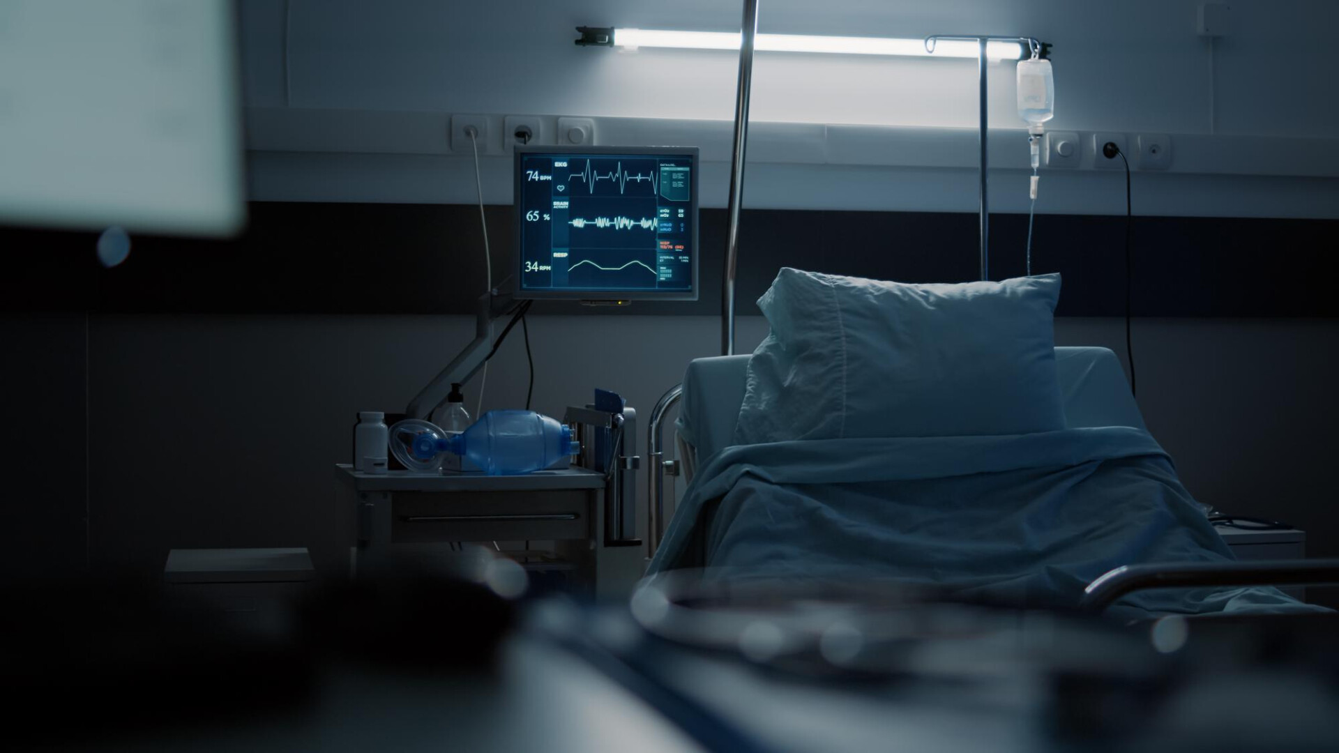 Hospital ICU bed with monitor showing vital signs, IV drip, and assorted equipment in a dimly lit room.