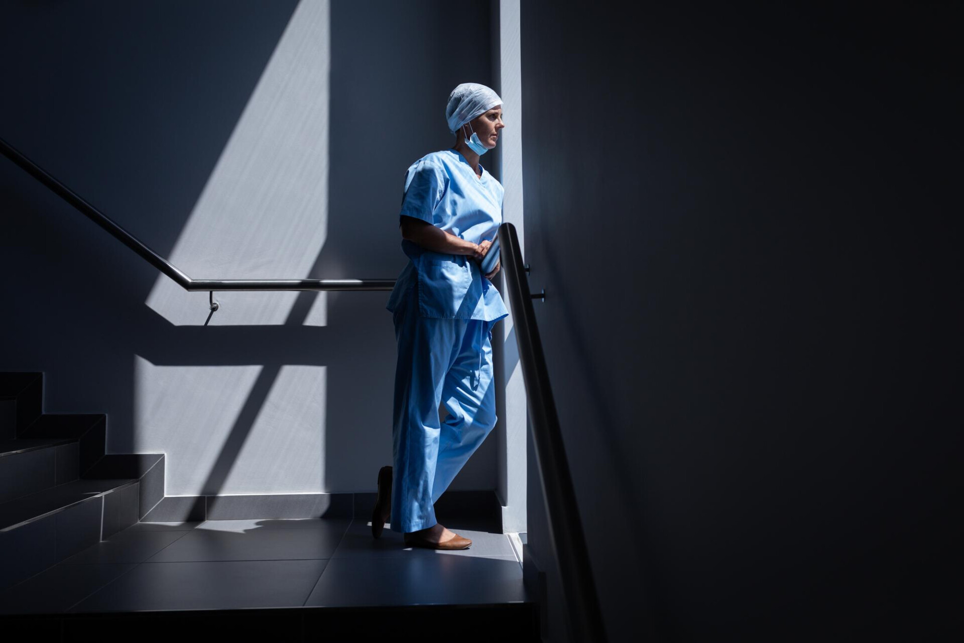 Healthcare worker in blue scrubs and cap stands on a stair landing, mask lowered under the chin, in a dimly lit hallway with dramatic shadows, looking ahead
