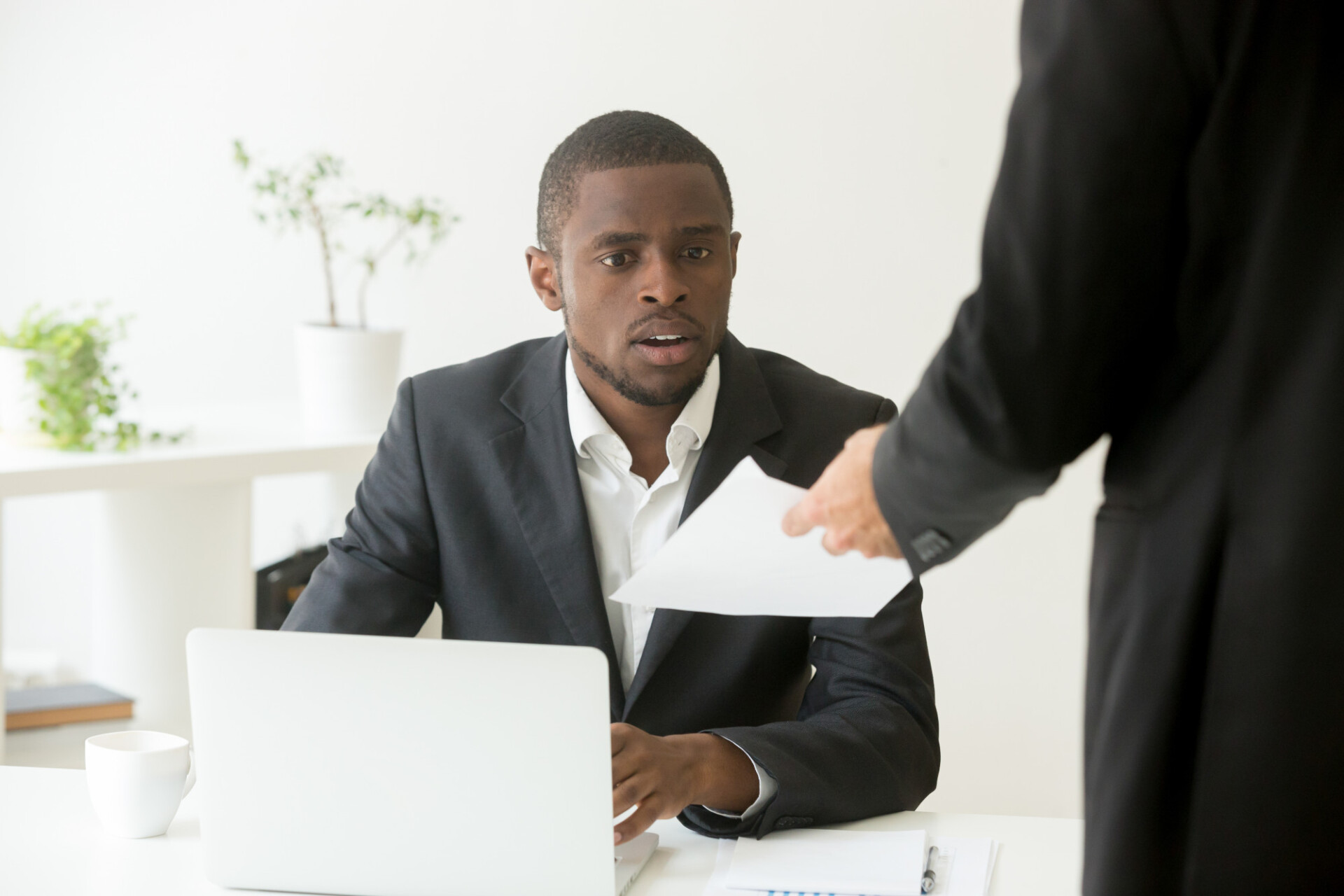 Man in a suit at a white desk receives a document from a standing colleague.