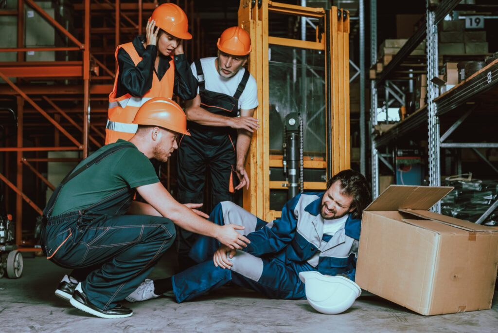 How To Claim Compensation For A Workplace Injury In The UK - Wolf Law Wolf Law 677 Group of workers in orange helmets helping an injured colleague on the warehouse floor, surrounded by boxes and shelving.