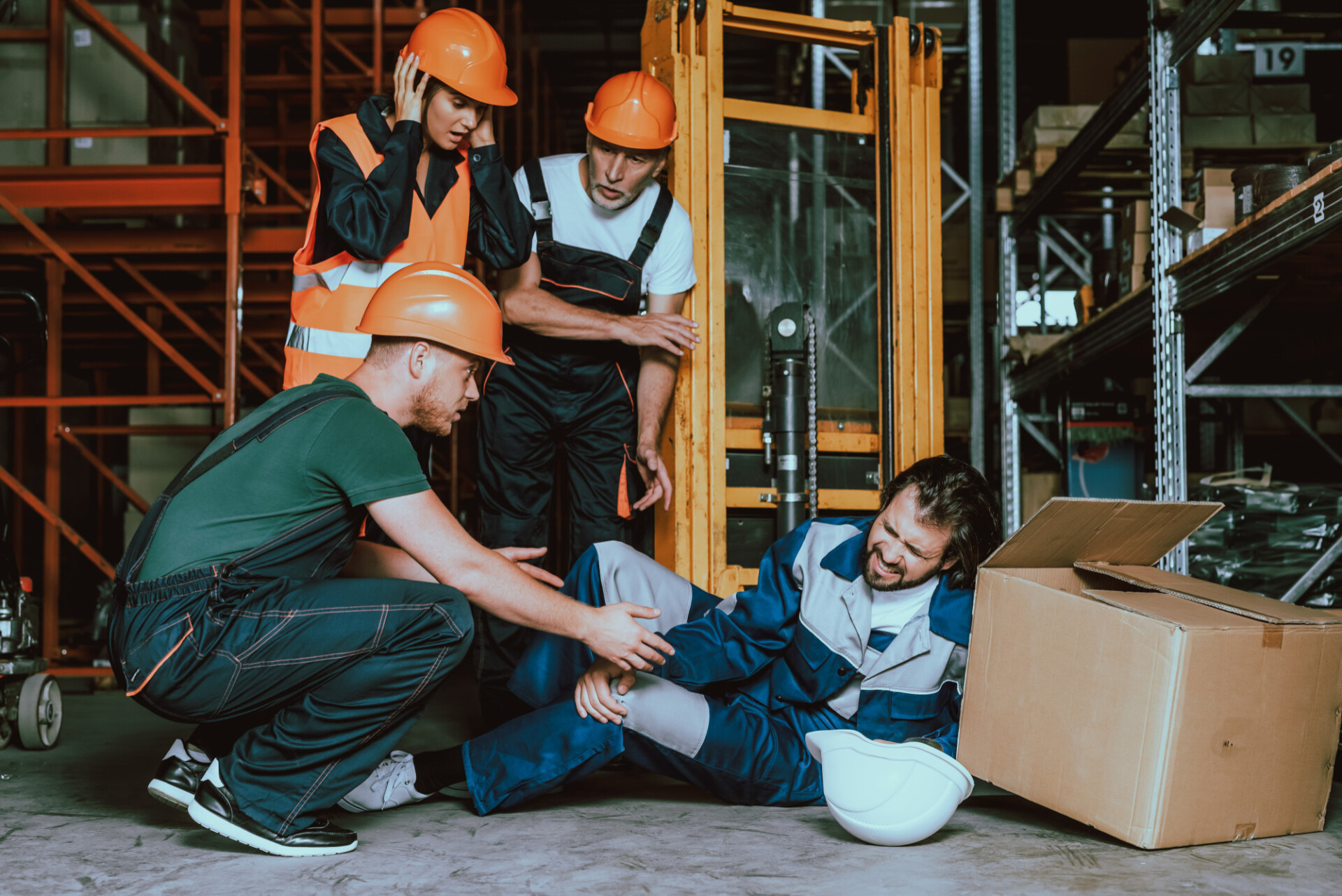 Group of workers in orange helmets helping an injured colleague on the warehouse floor, surrounded by boxes and shelving.