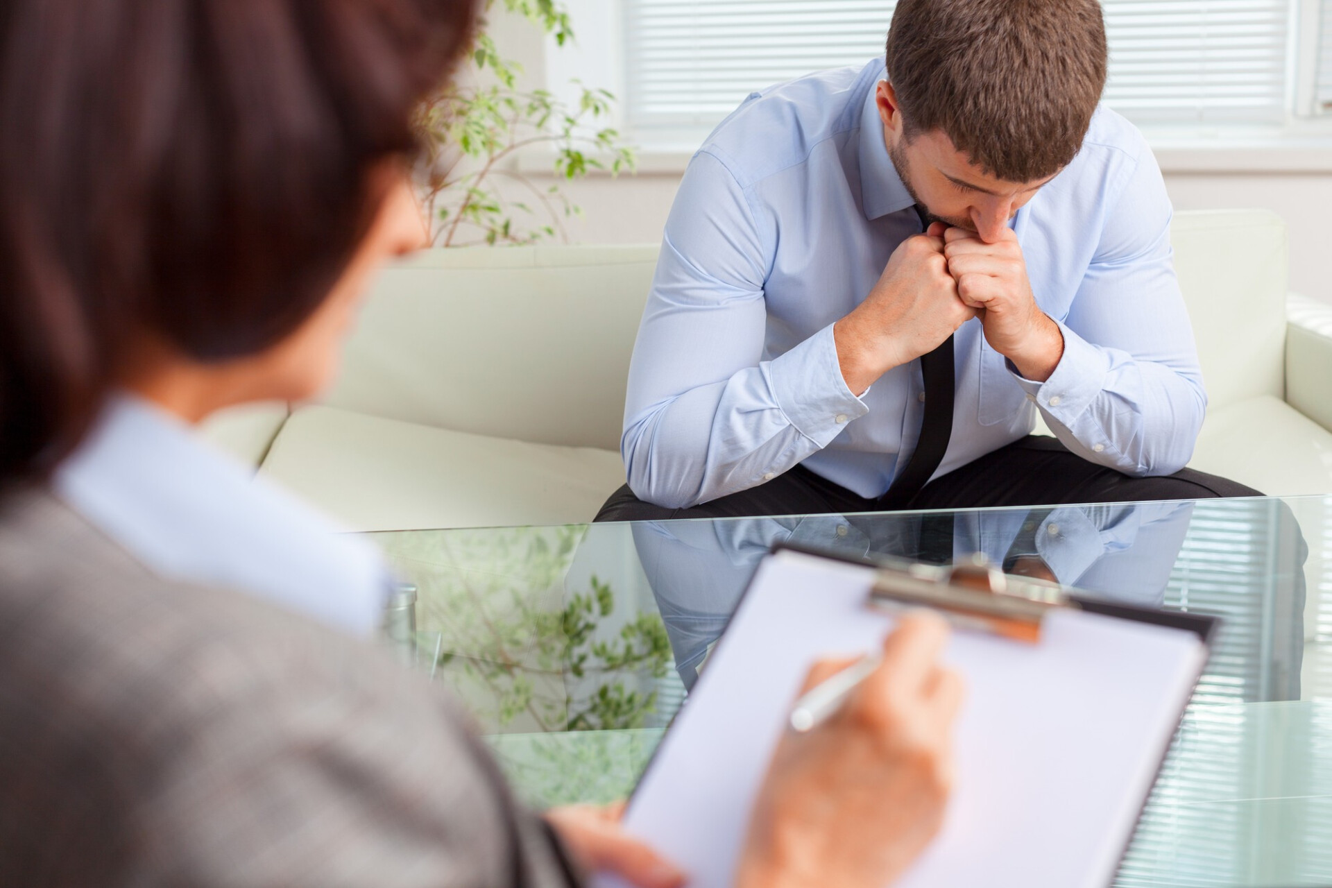 Man in a light blue shirt sits on a couch, elbows on his knees, hands clasped under his chin, looking down in a therapy session.