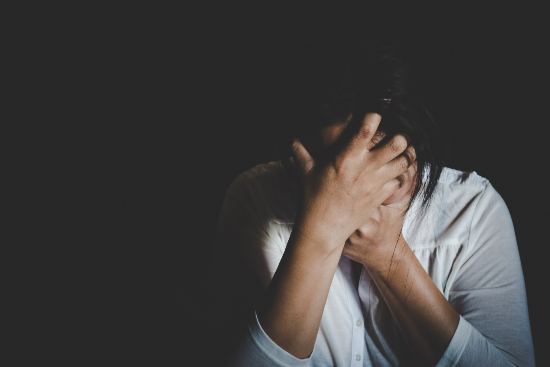 Person sitting in a dark room, hands pressed to their head in distress or frustration.