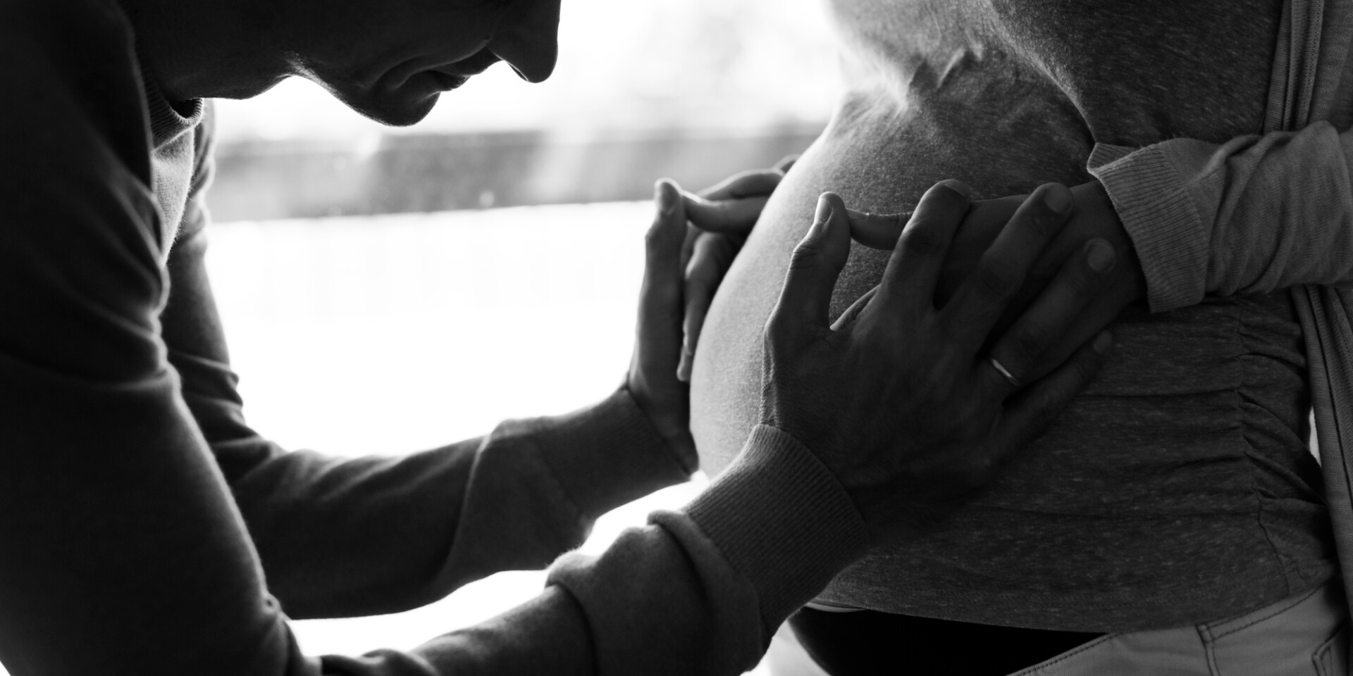 A person and partner gently cradle a pregnant belly with intertwined hands in profile view (black and white).
