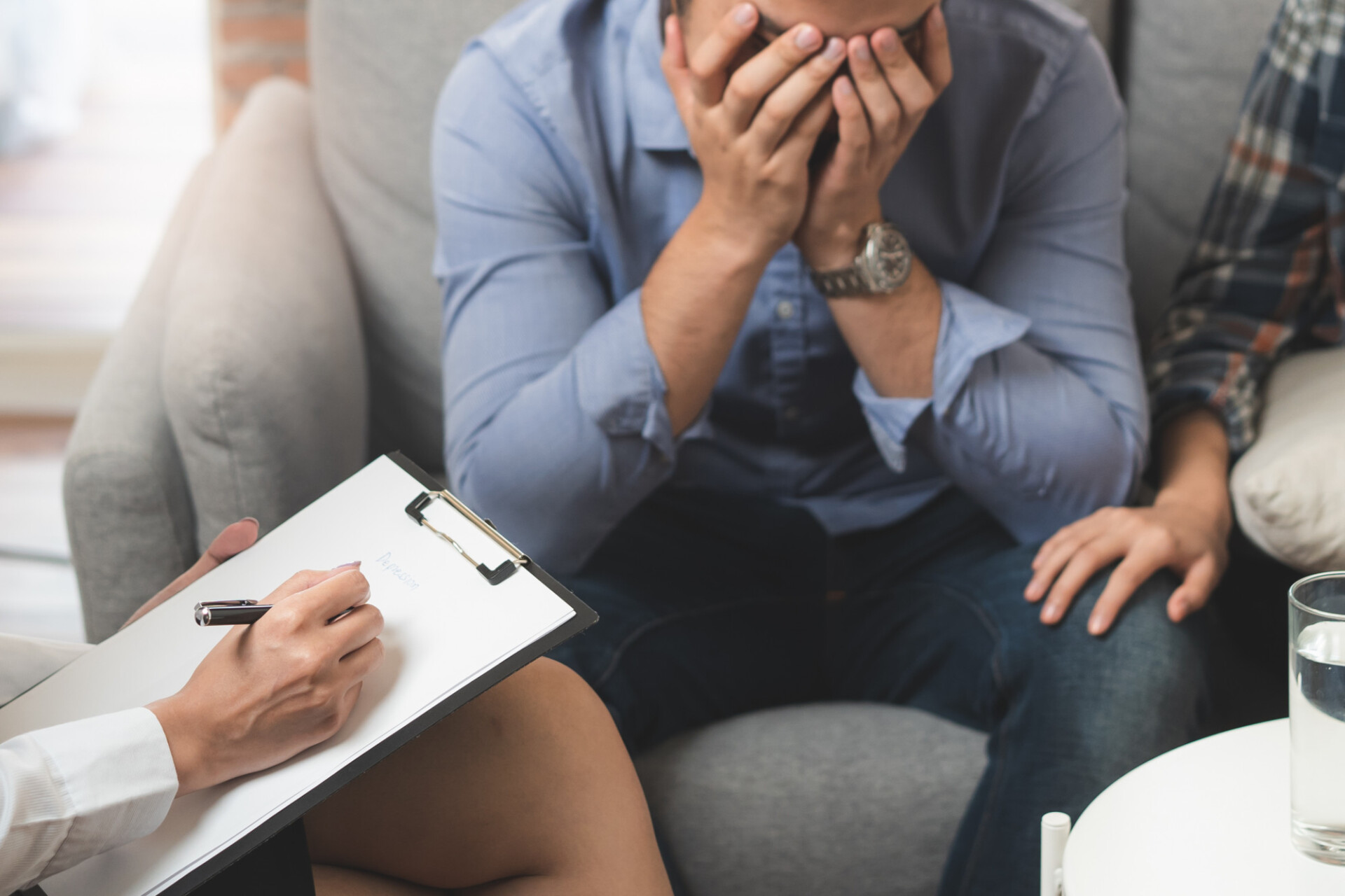 Person sits on a couch, hands over face in distress during a counseling session while another person writes on a clipboard nearby