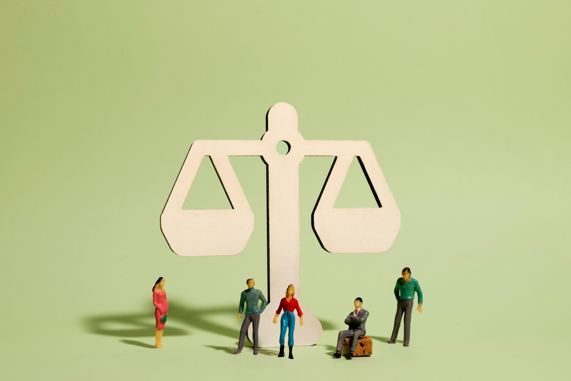 Miniature people stand around a large white balance scale, symbolizing fairness and weighing decisions on a green background.