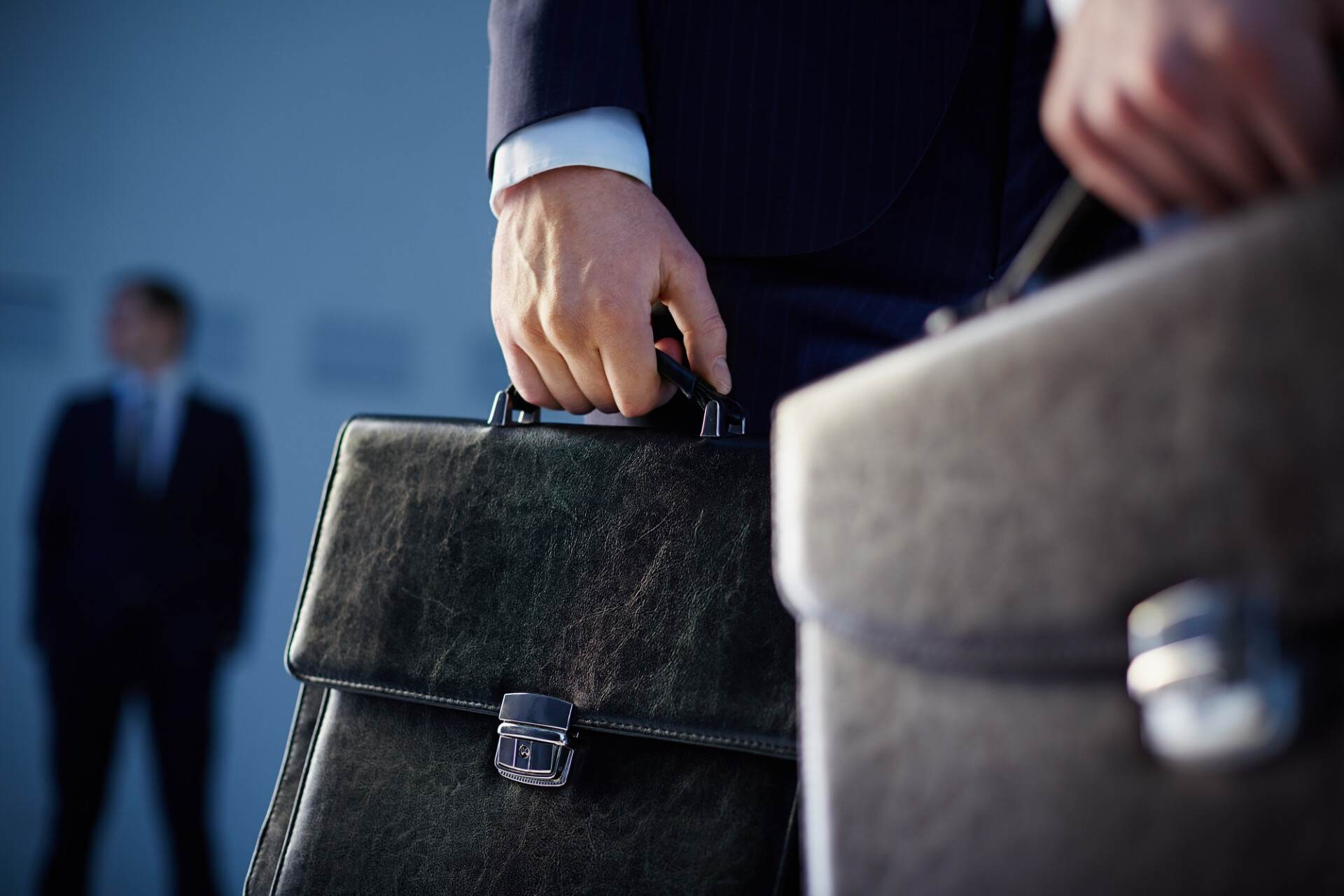 Close-up of a hand gripping a black leather briefcase, with a suited person in the background.