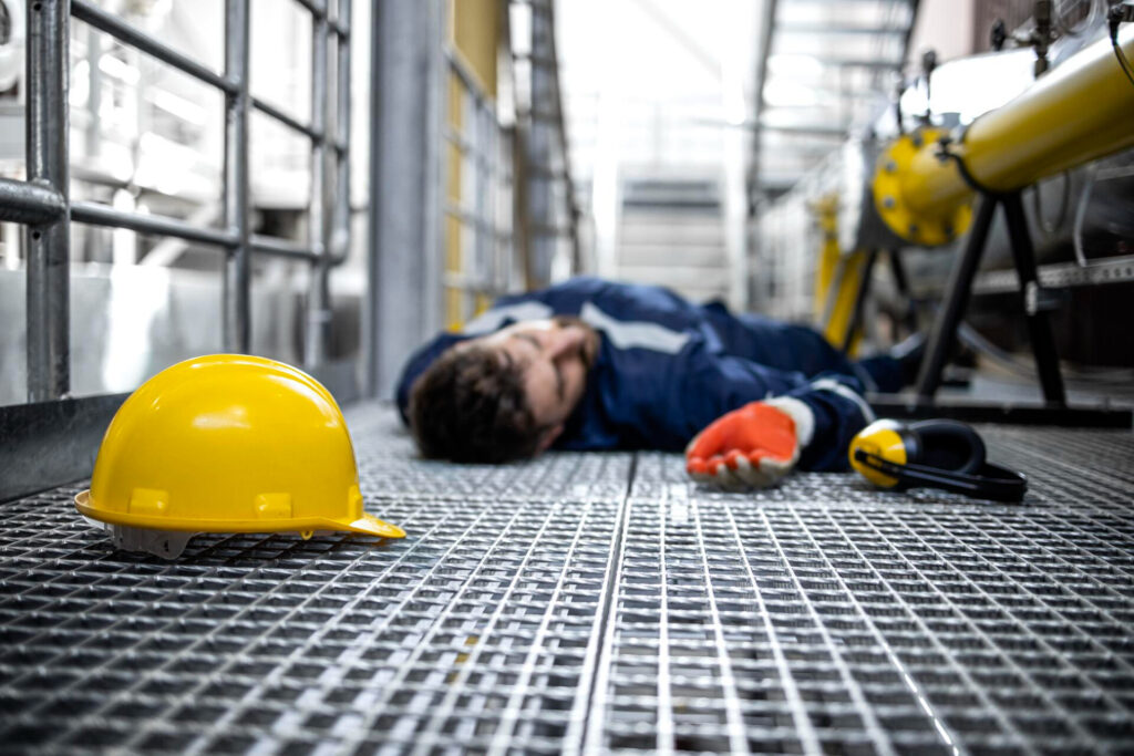Leg amputation after a workplace accident: what can an injured worker claim? - Wolf Law Wolf Law 3817 Worker lies on metal grate floor in industrial facility, yellow safety helmet in foreground and scattered PPE nearby indicating an accident or injury.