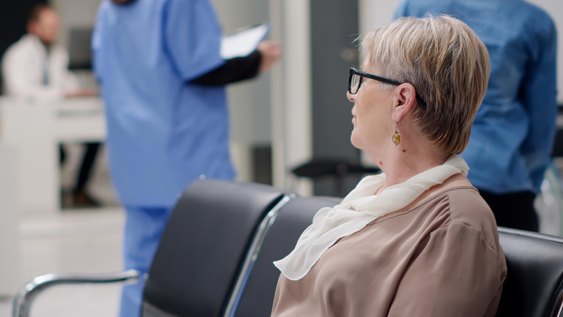 Older woman with glasses and a white scarf sitting in a medical clinic waiting area; staff in blue scrubs blurred in the background.