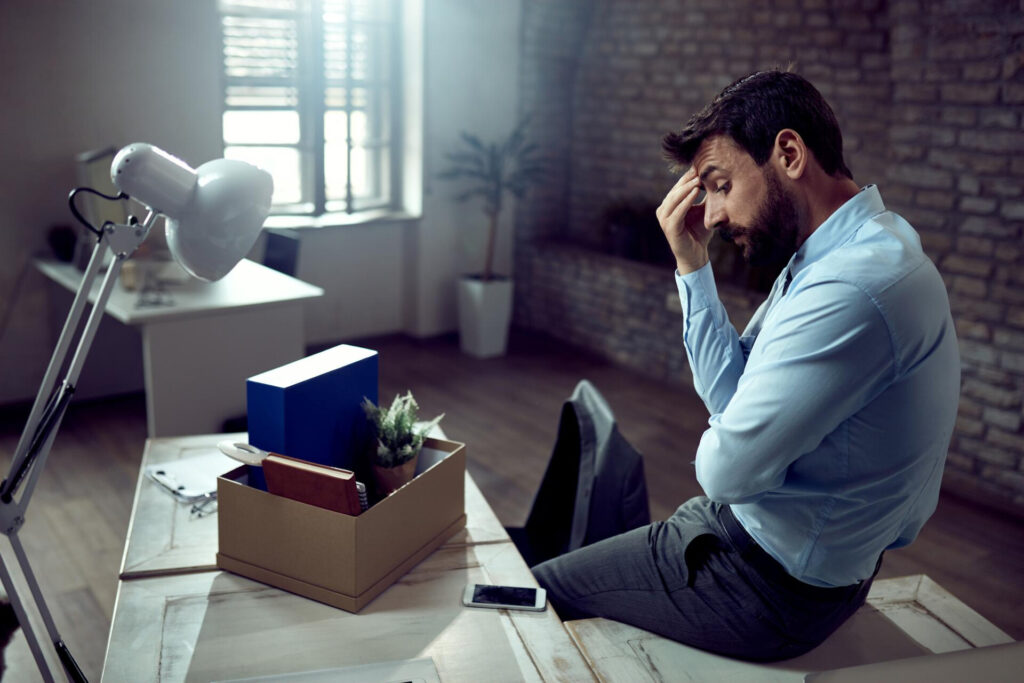 Removal of the unfair dismissal cap: what employers and workers need to know - Wolf Law Wolf Law 6354 Overwhelmed man in a blue shirt sits at a desk with a cardboard box of items, hand on his forehead in a bright office.