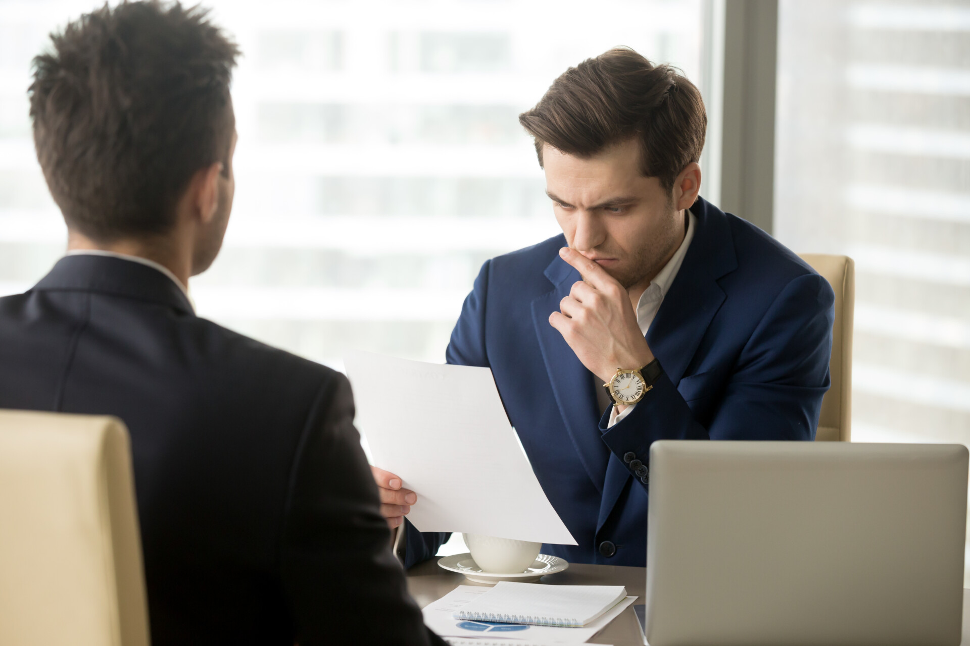 Two businessmen in a meeting: one in a blue suit studying a document with a thoughtful pose, the other facing away with his back to the camera, a laptop on the table and a coffee cup nearby.