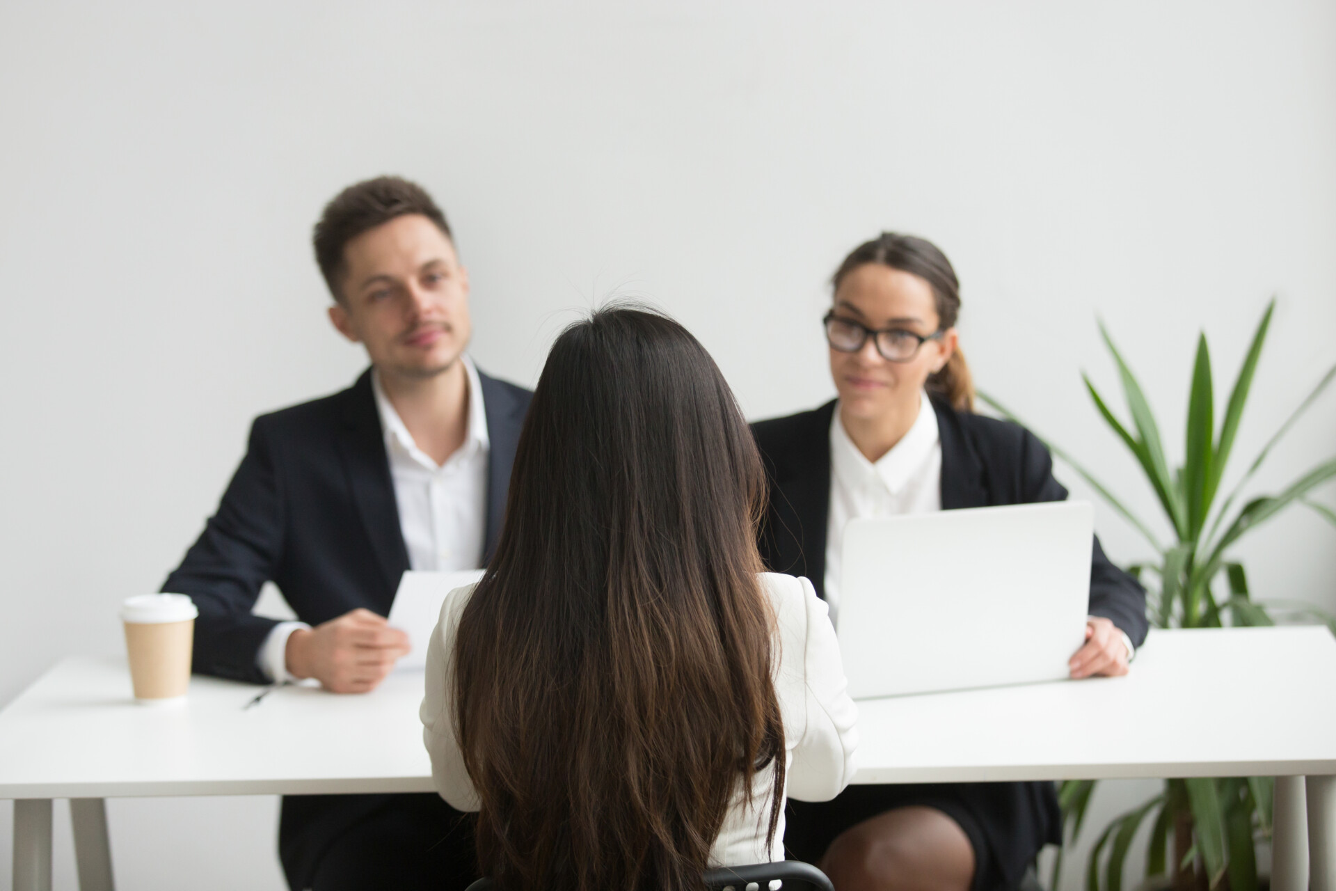 Woman with long dark hair seen from behind sits at a white table facing two colleagues in business attire across from her; a plant is nearby in a bright office.