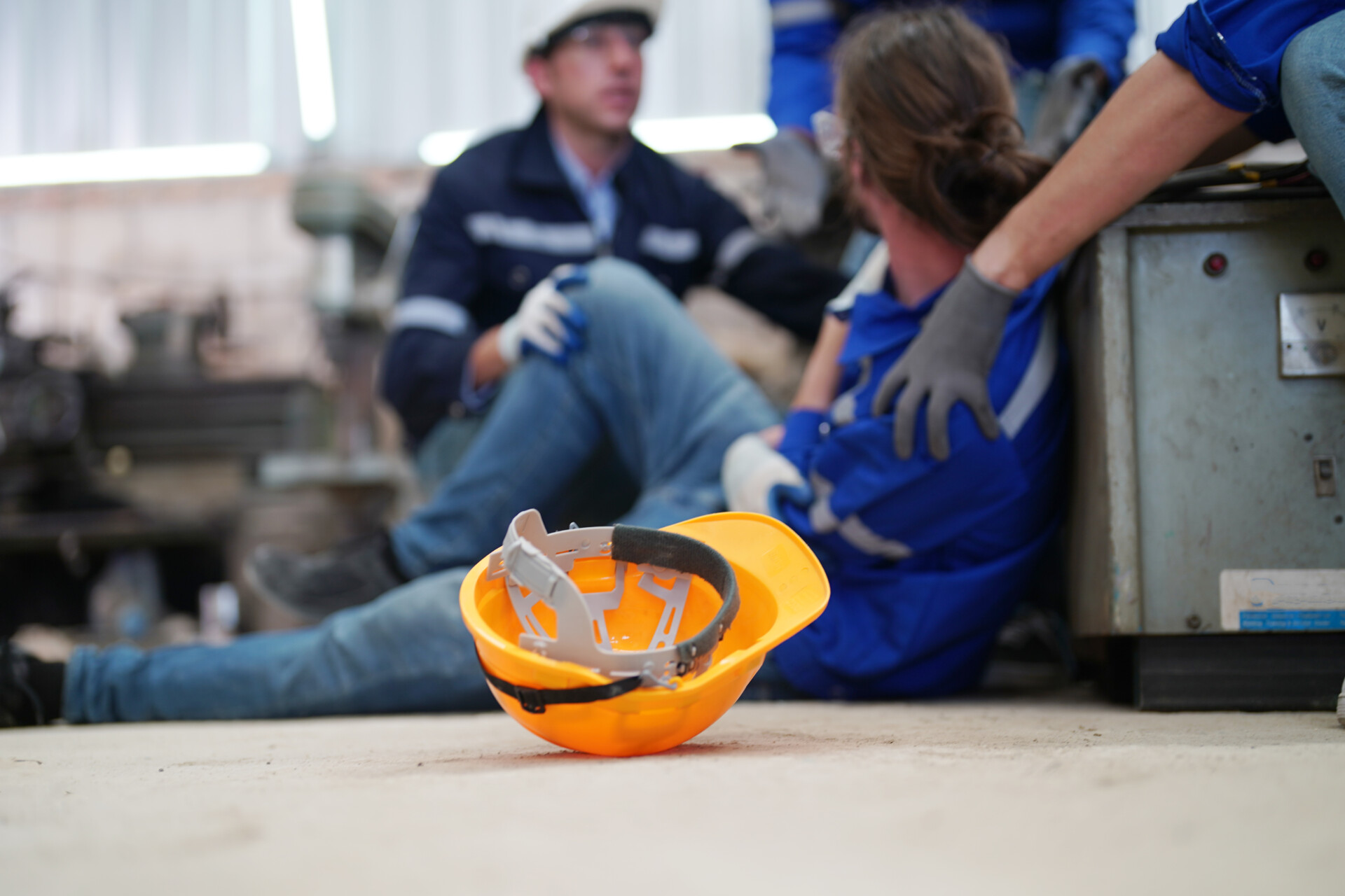 Orange safety helmet on the floor in the foreground, with two workers resting and talking in a blue-workwear shop in the blurred background.