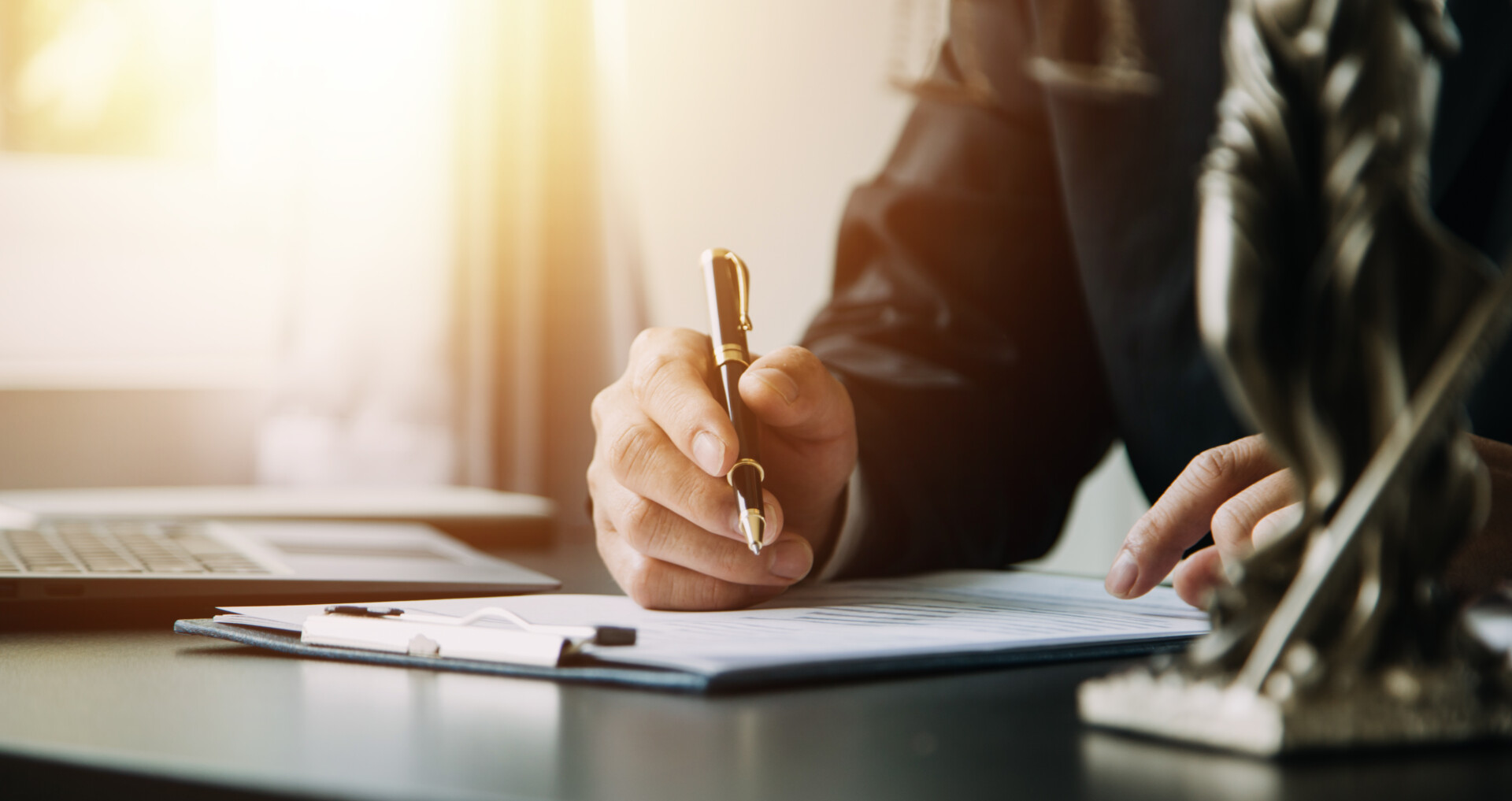 Professional signs documents at a desk, pen in hand over papers with a laptop nearby, sunlight streaming in.