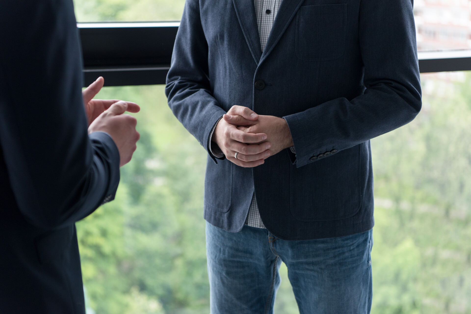 Two people in dark blazers speak with hands clasped in front of a window; view focused on the hands and torsos outdoors.