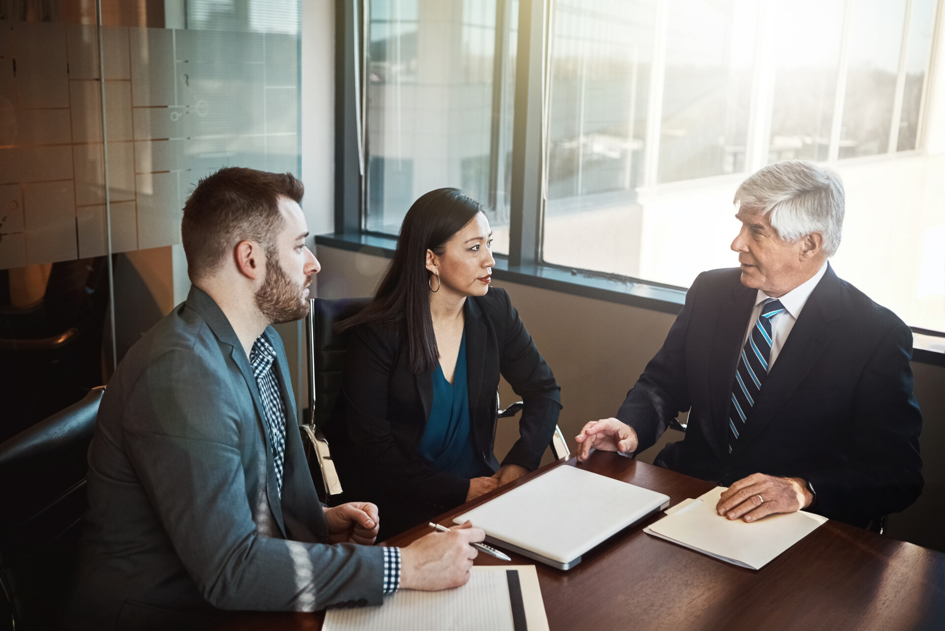 Three professionals sit around a conference table discussing documents and a laptop in a bright office setting