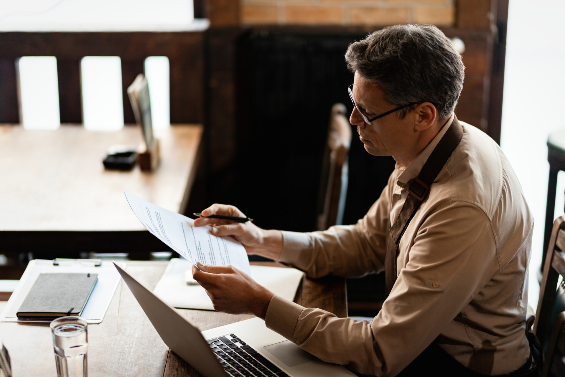 Man in a beige shirt and glasses reads a document at a wooden desk with a laptop nearby.