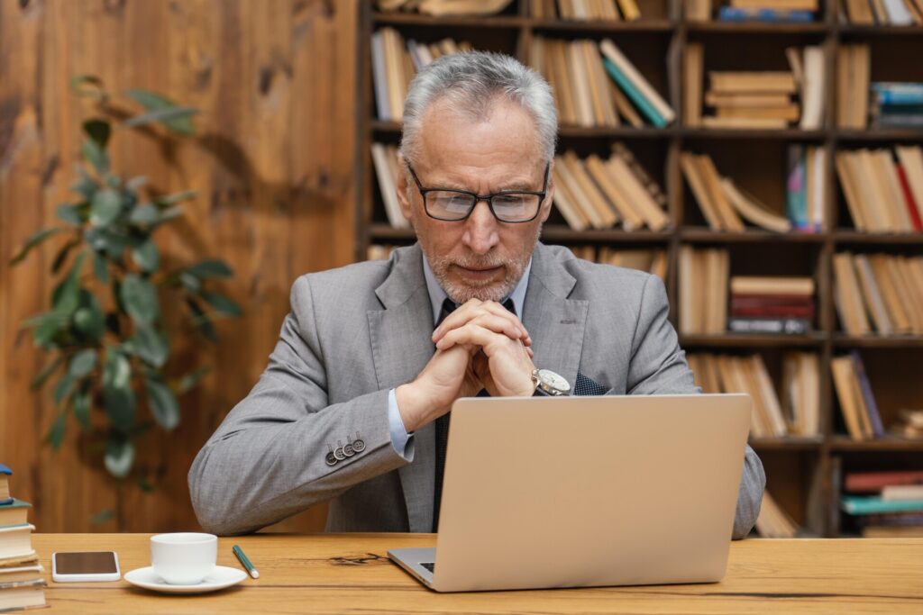 NEW ASSOCIATE APPOINTMENTS AT SCOTT REES & CO - Wolf Law Wolf Law portrait-elegant-old-man Older man in glasses and a gray suit sits at a wooden desk with a laptop, hands clasped in front, in a library.